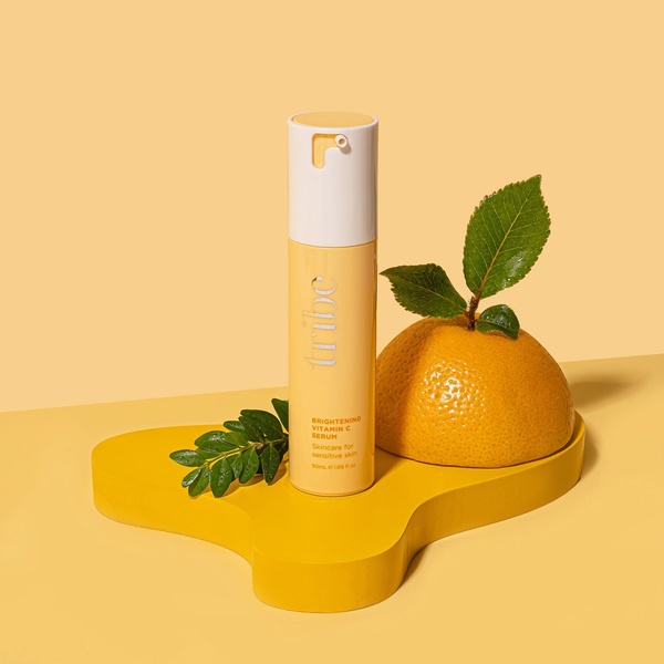 A clean, high-contrast product hero shot featuring a Vitamin C serum bottle centered on a sculptural yellow platform with natural citrus elements.